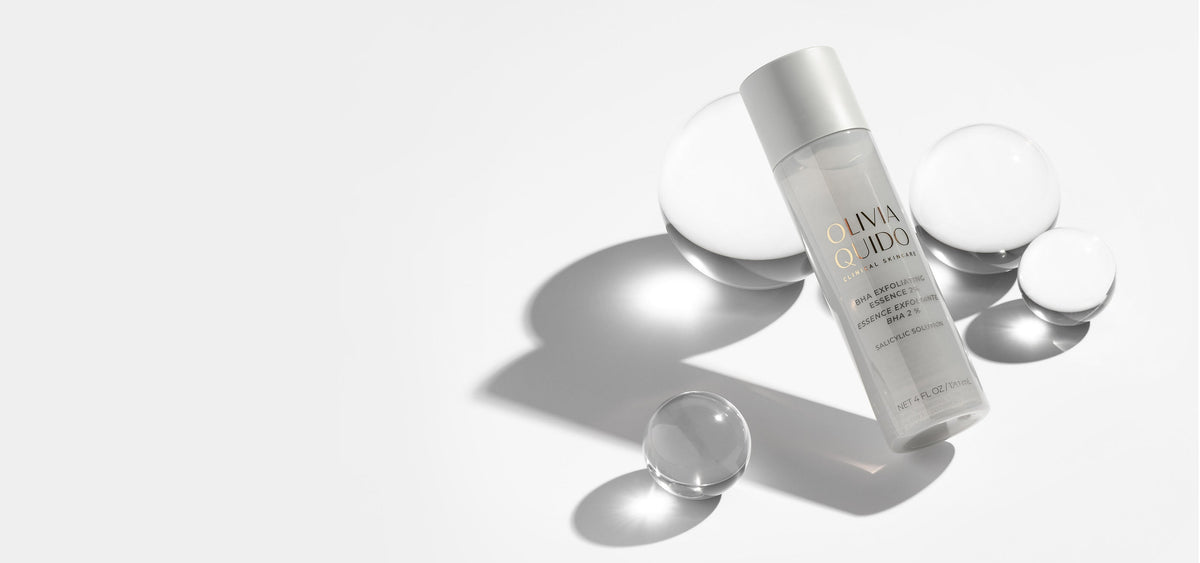 Olivia Quido BHA Exfoliating Essence bottle leaning on glass sphere in grey environment